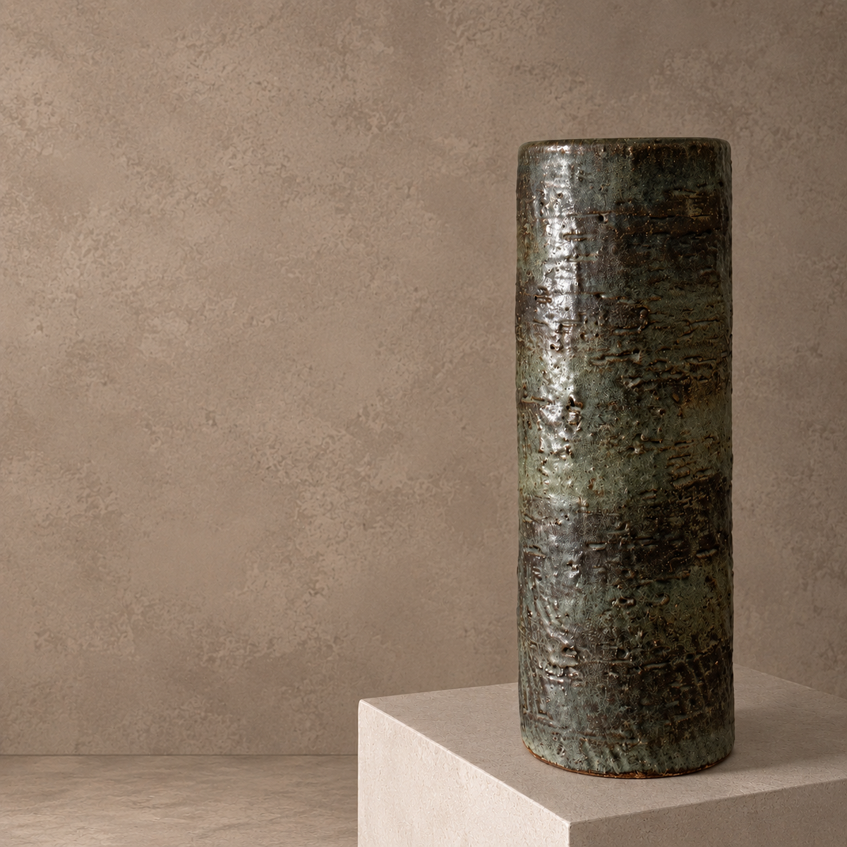 A tall cylindrical ceramic vase with a textured, metallic bronze and green glaze, displayed on a light beige pedestal against a textured beige wall.