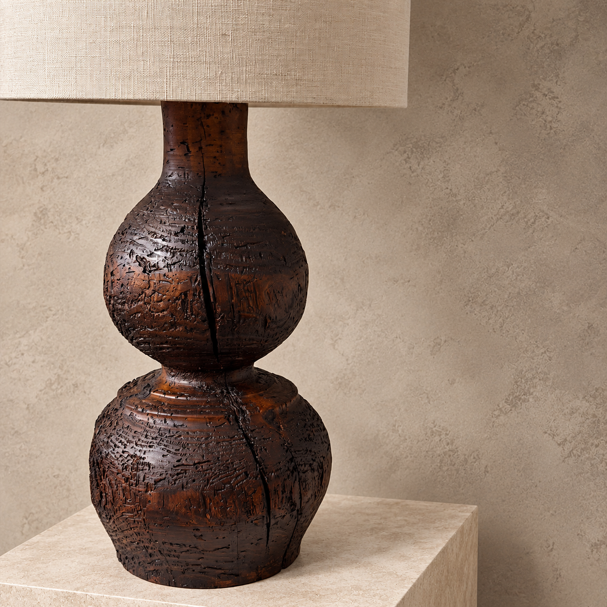 A rustic dark brown wooden table lamp with a textured, crackled finish and a beige linen drum shade, displayed on a stone pedestal against a textured beige wall.