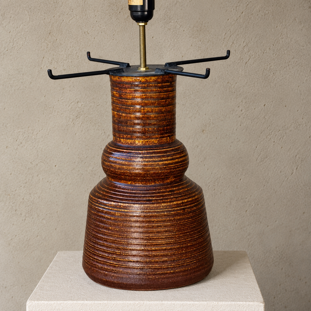 A ceramic table lamp base with a textured, ribbed brown and amber glaze, featuring a black metal crossbar shade holder and a brass socket, displayed on a light beige pedestal against a neutral textured wall.