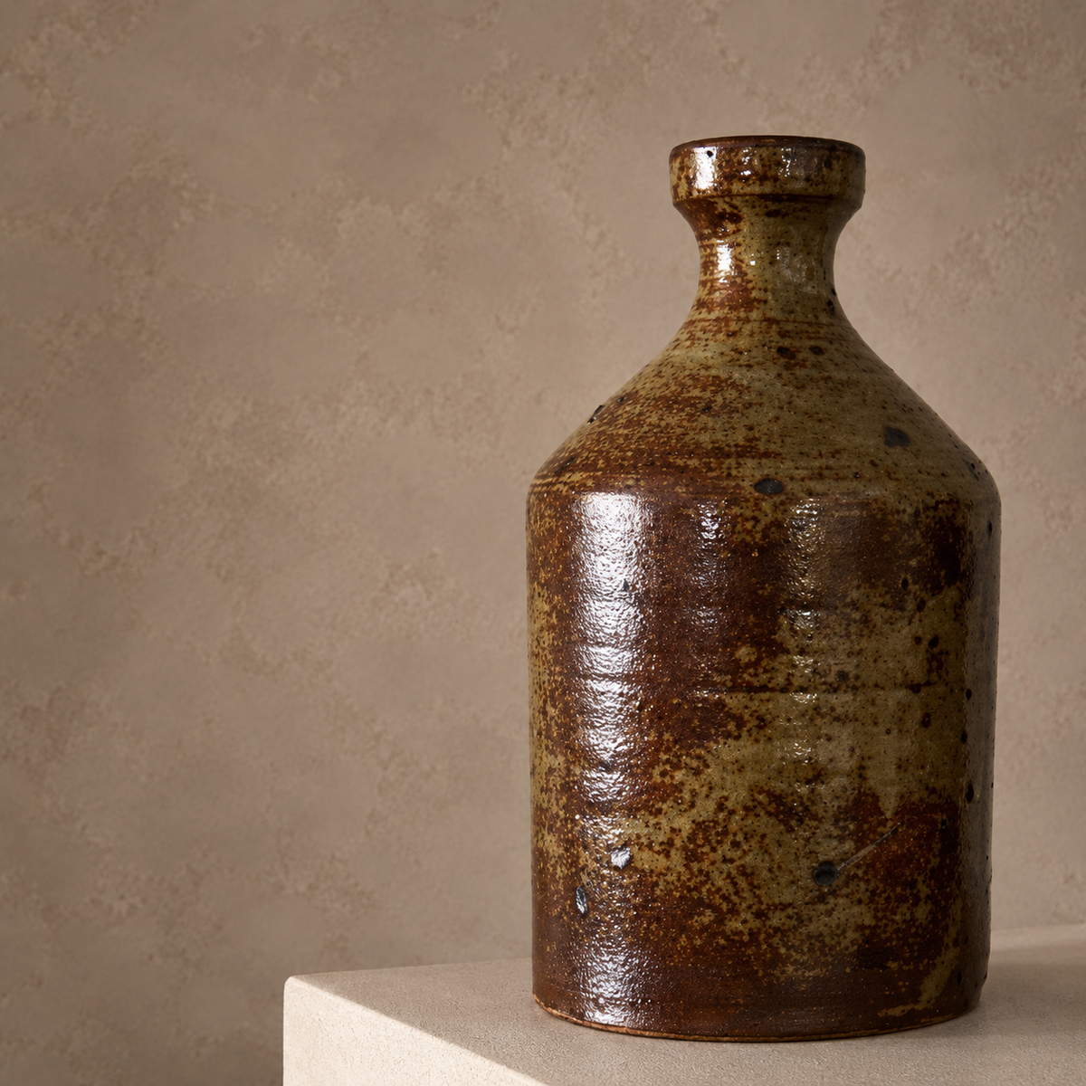 A rustic ceramic bottle vase with a textured, glossy brown and olive-green glaze, displayed on a light beige pedestal against a neutral textured beige wall.