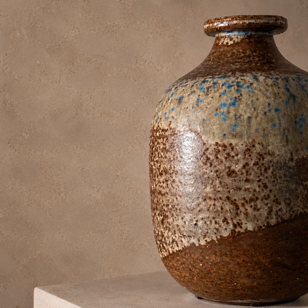 A close-up of a ceramic vase with a rustic, hand-painted glaze featuring brown, beige, and blue speckled patterns, displayed on a light stone pedestal against a textured beige wall.