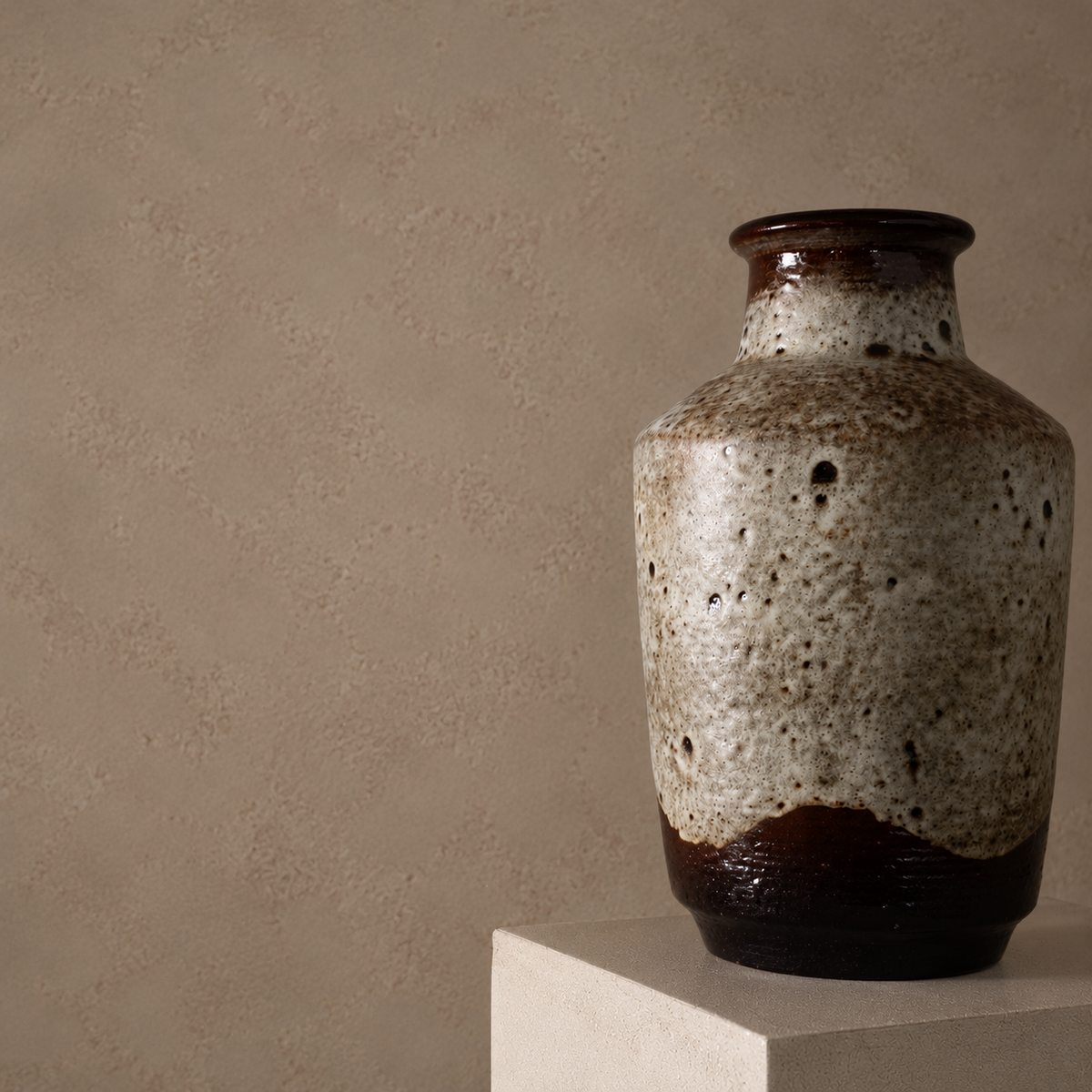 A rustic ceramic vase with a speckled beige and brown glaze and a dark brown base, displayed on a white pedestal against a textured beige wall.