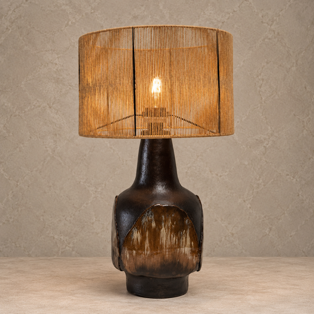 A table lamp with a dark brown textured ceramic base and a cylindrical woven bamboo shade, lit with a warm Edison bulb against a neutral textured beige wall.
