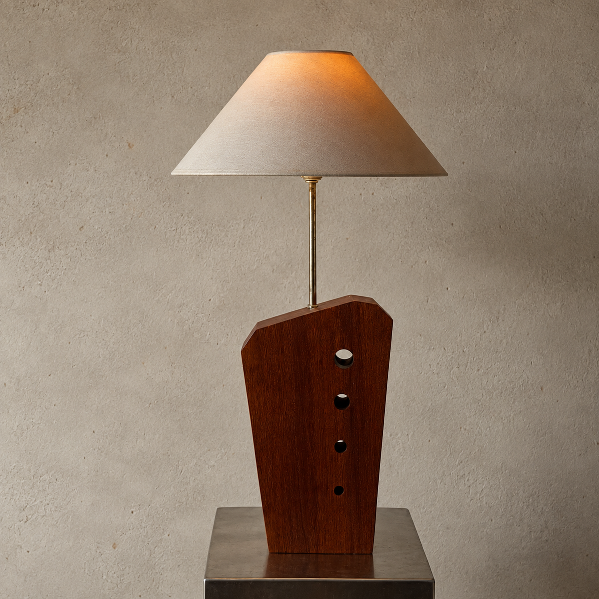 A modern table lamp with a dark brown wooden geometric base, a slim brass stem, and a beige fabric shade glowing with warm light, set on a metallic pedestal against a textured beige wall.
