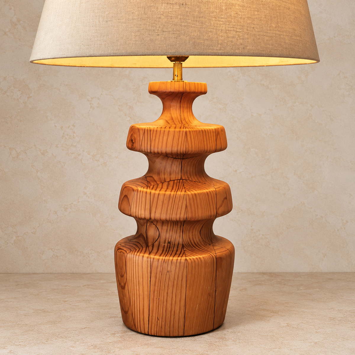 A table lamp with a sculptural, tiered wooden base in a warm honey-brown finish, topped with a beige linen drum shade and a brass neck, set against a textured beige wall and floor.