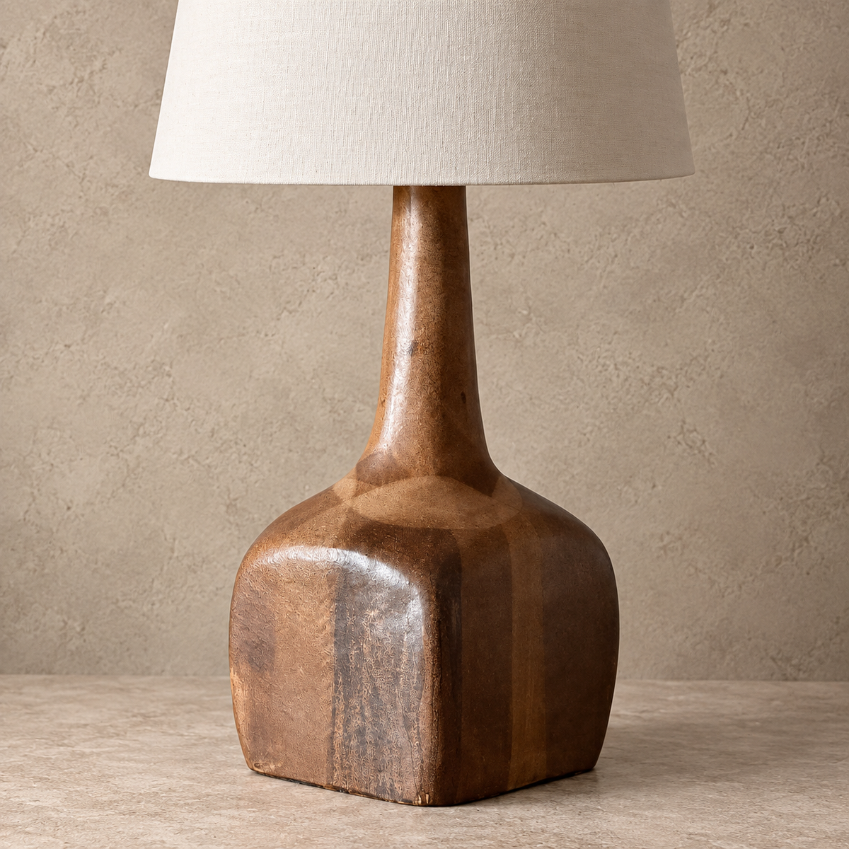 A sculptural table lamp with a polished brown wooden base and a tapered off-beige fabric shade, set against a textured beige wall and stone surface.