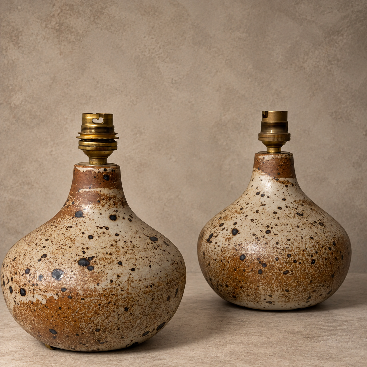 Two handmade ceramic table lamps with rounded bulb-shaped bodies, speckled brown and beige glaze, and brass lamp sockets, displayed on a white shelf against a textured gray wall.