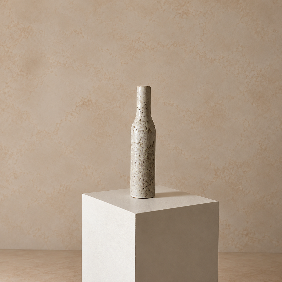 A tall, slender ceramic vase with a speckled beige and gray glaze stands centered on a white pedestal against a textured beige wall.