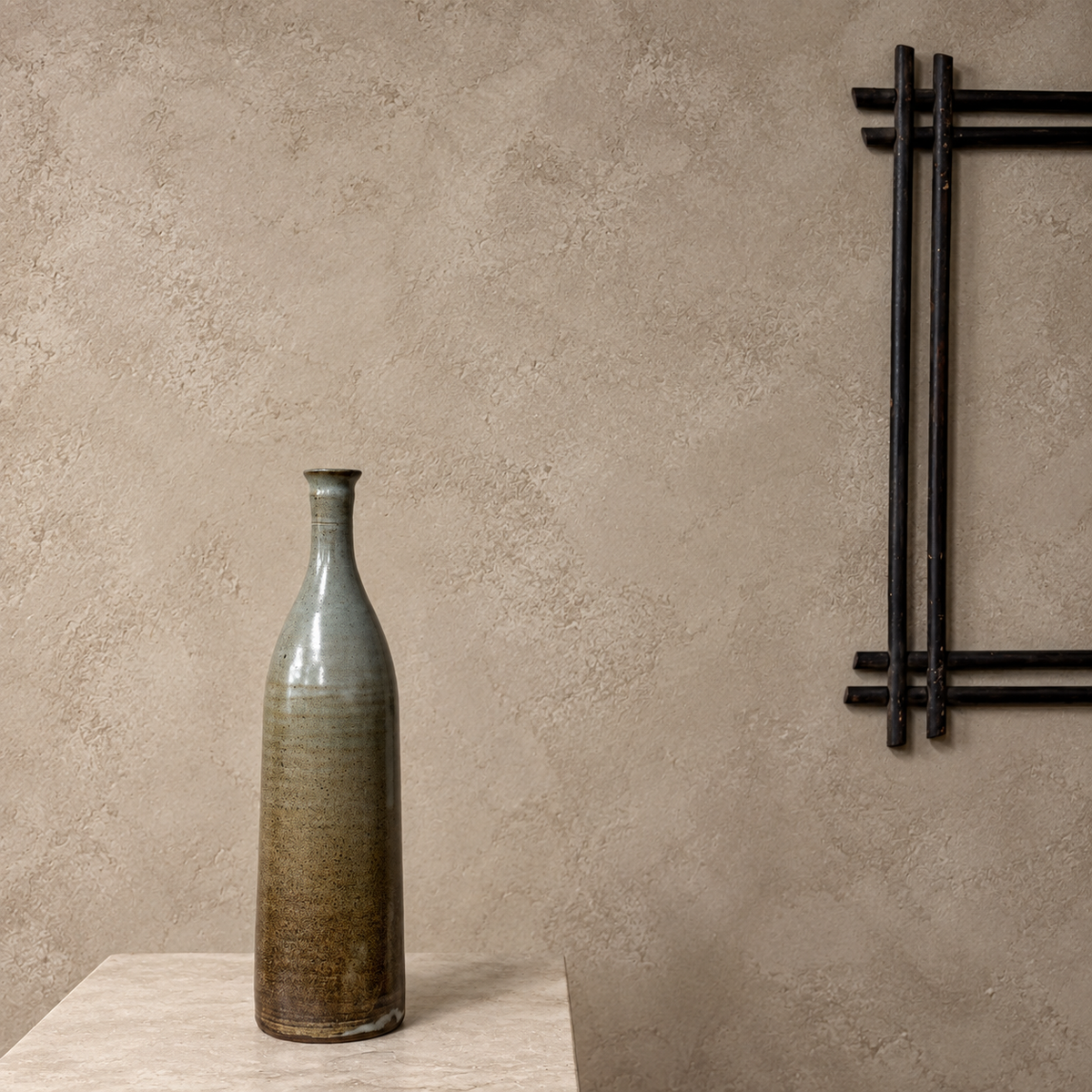 A tall, narrow ceramic vase with a textured glaze transitioning from gray-green at the top to earthy brown at the base, displayed on a white pedestal against a textured concrete wall.