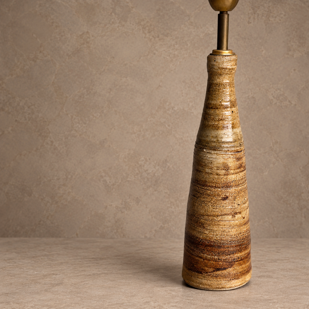 A ceramic table lamp base with a tapered, textured cone shape in earthy brown and beige tones, topped with a brass finial and stem against a neutral textured beige background.