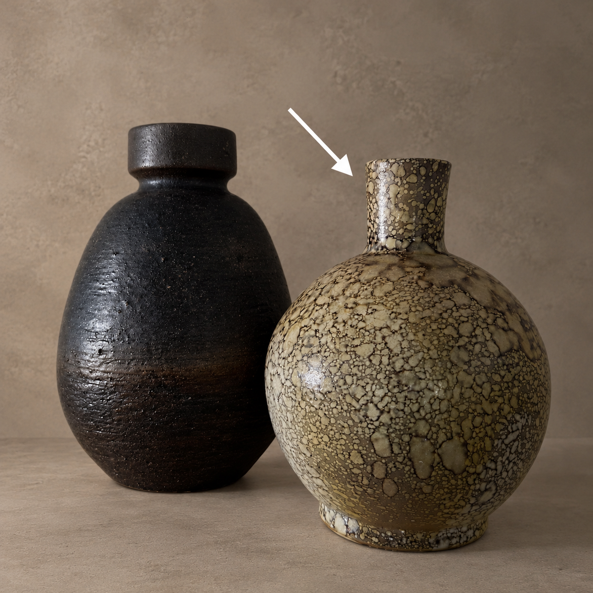 Two ceramic vases on a tabletop against a textured beige wall. The left vase is matte black with a rounded body and short neck.