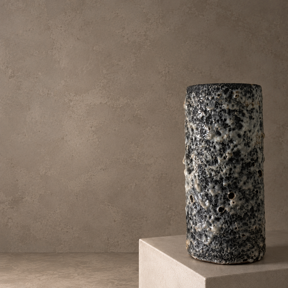 A tall cylindrical ceramic vase with a textured, porous black and white speckled glaze, displayed on a light beige pedestal against a neutral concrete-textured background.