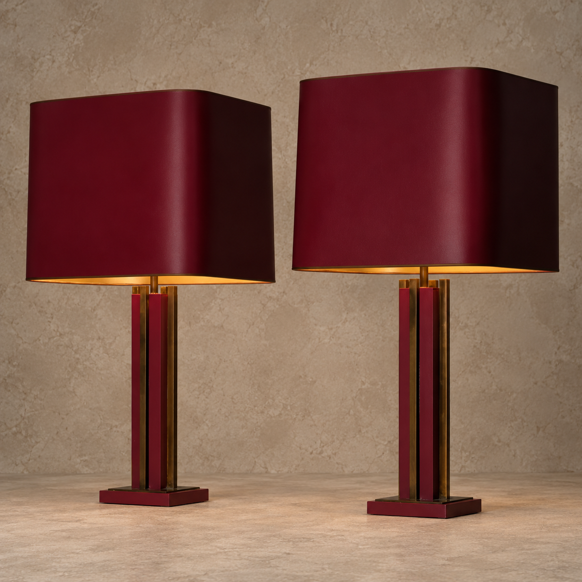 A pair of modern table lamps with dark burgundy shades and matching burgundy bases accented by vertical gold-toned metal bars, set against a textured beige wall and floor.
