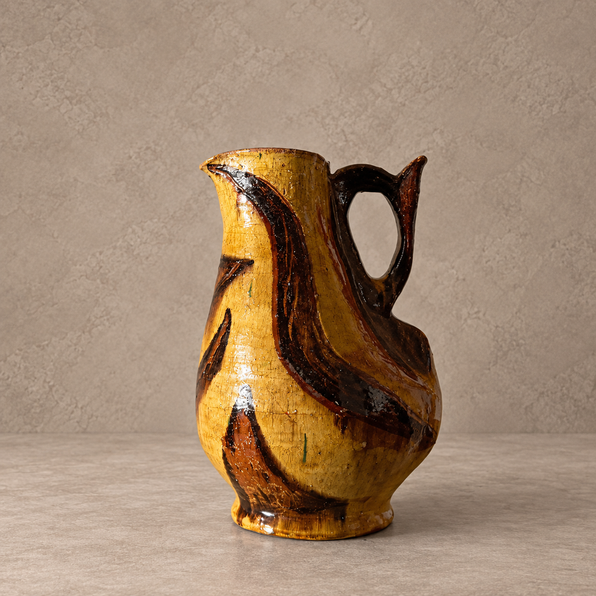 A handmade ceramic pitcher with a glossy yellow and dark brown abstract swirled glaze, set against a neutral textured beige background.