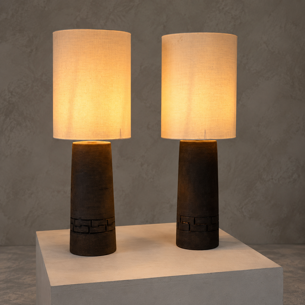 Two matching table lamps with cylindrical warm-white fabric shades and dark brown carved wooden bases, displayed side by side on a light pedestal against a textured gray wall.