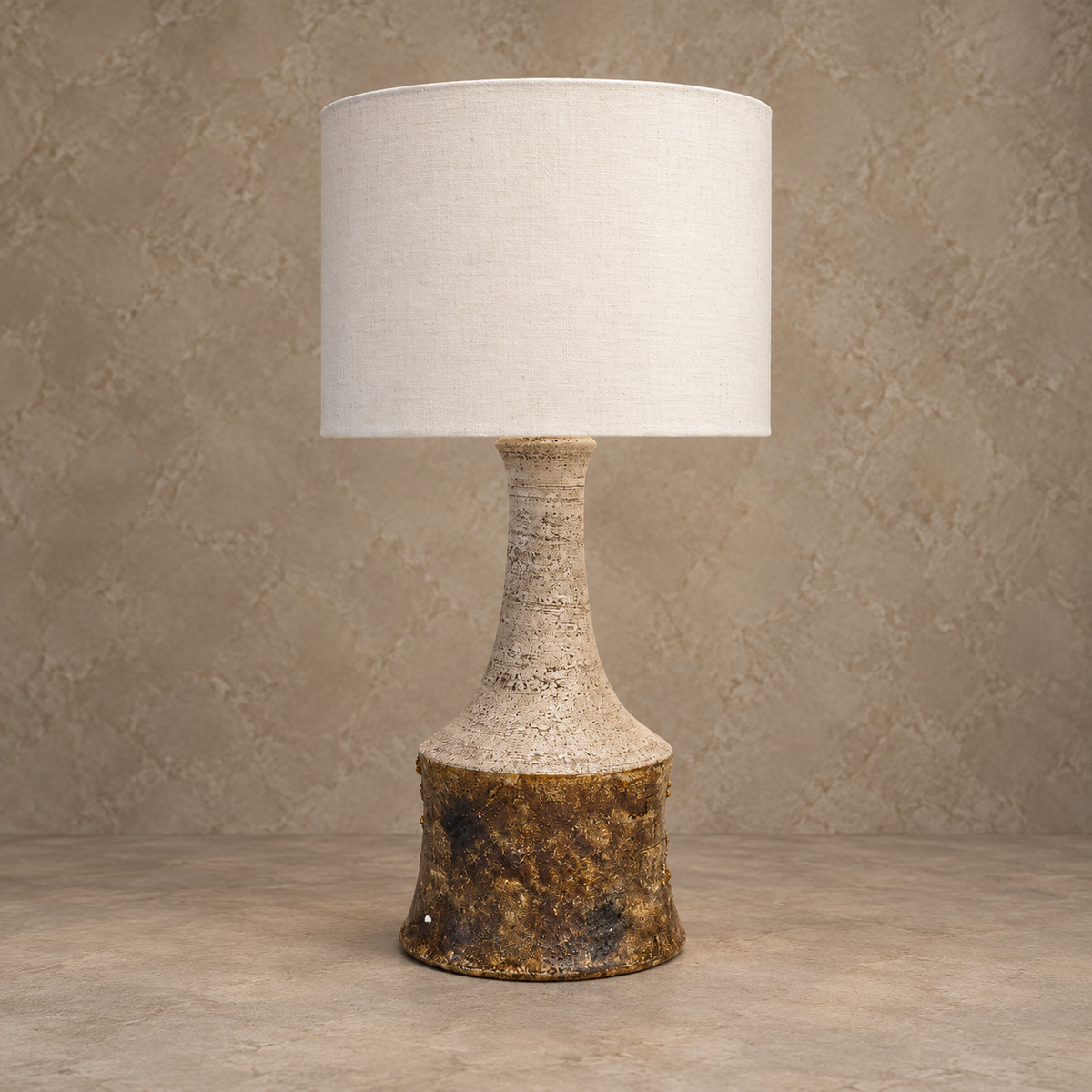 A table lamp with a textured, tapered ceramic base featuring a mottled gold and brown finish, paired with a large, cylindrical off-white fabric drum shade, set against a neutral, textured beige wall and floor.