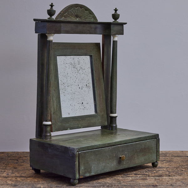 A green-painted dresser mirror with a tilt mirror, Ionic columns, and a drawer with a brass handle, set against a plain background.